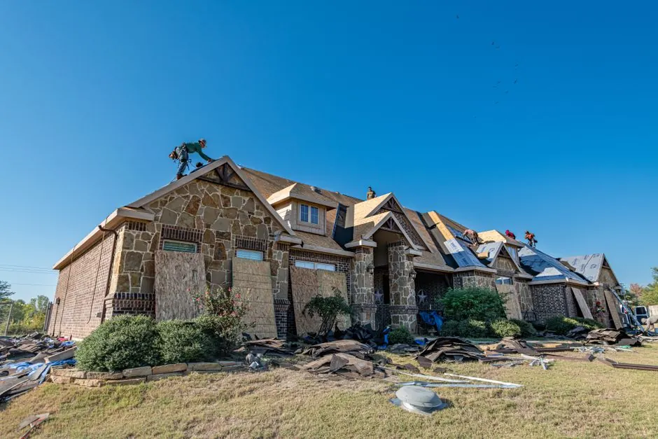 Brick home undergoing professional roof installation with workers on the roof under a clear sky.